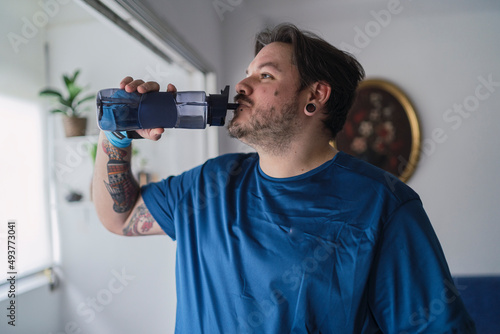 hombre con camiseta azul y tatuajes en el brazo bebiendo agua. hombre de estilo alternativo bebiendo de una botella de plástico color azul