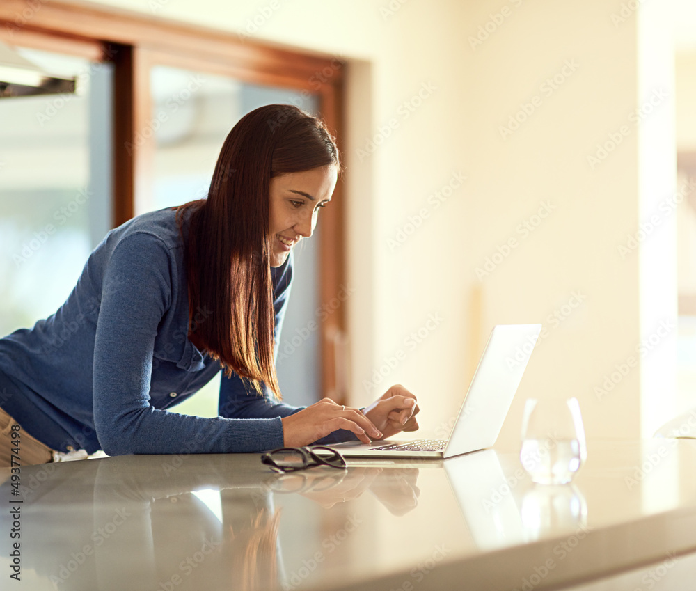 The kitchen is her hotspot. Shot of a happy young woman using her laptop while standing in her kitchen. The kitchen is her hotspot. Shot of a happy young woman using her laptop while standing in her kitchen.