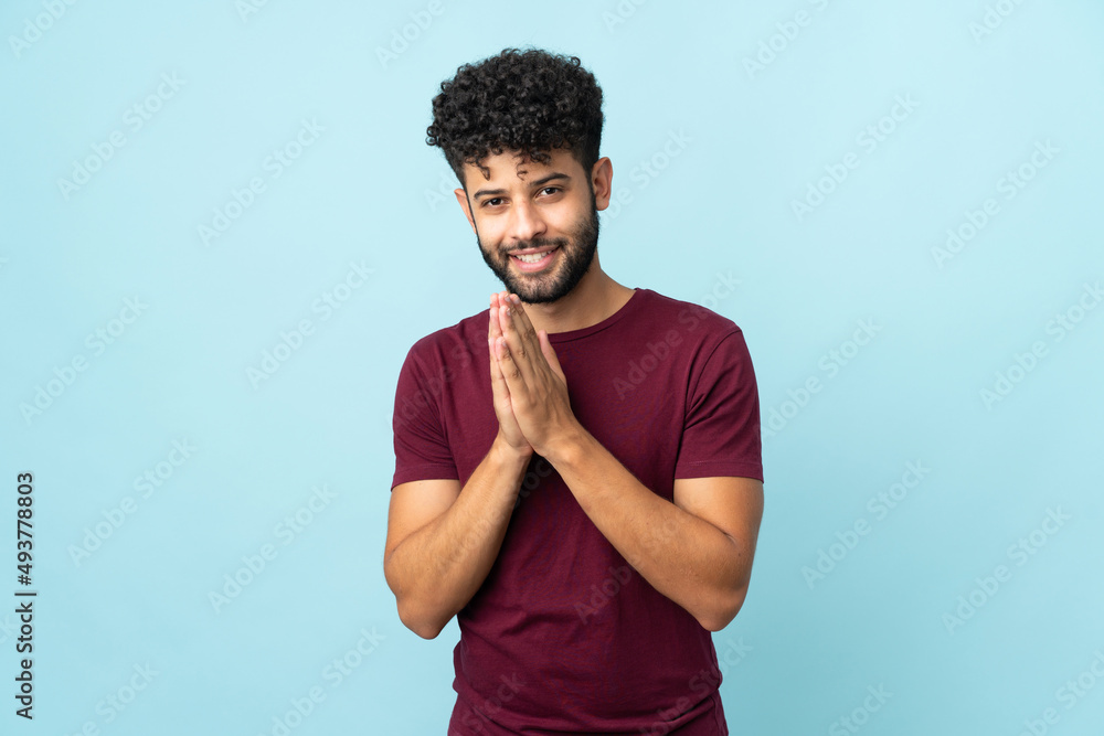 Young Moroccan man isolated on blue background keeps palm together. Person asks for something Young Moroccan man isolated on blue background keeps palm together. Person asks for something