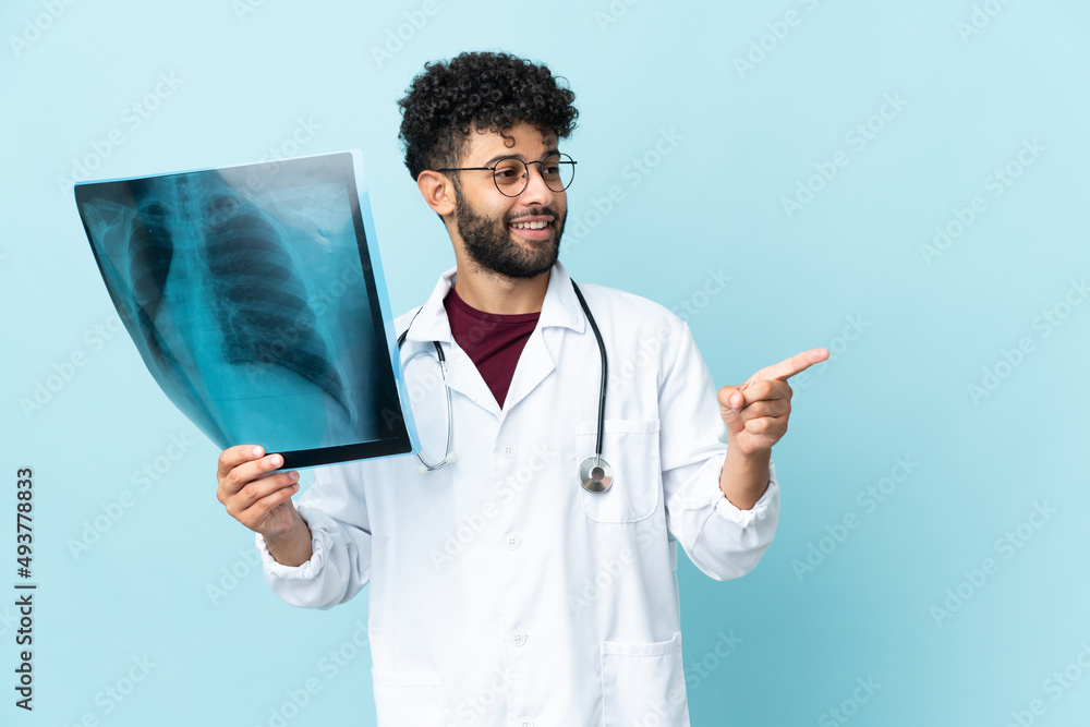 Young Moroccan traumatologist in workplace isolated on blue background pointing finger to the side and presenting a product Young Moroccan traumatologist in workplace isolated on blue background pointing finger to the side and presenting a product