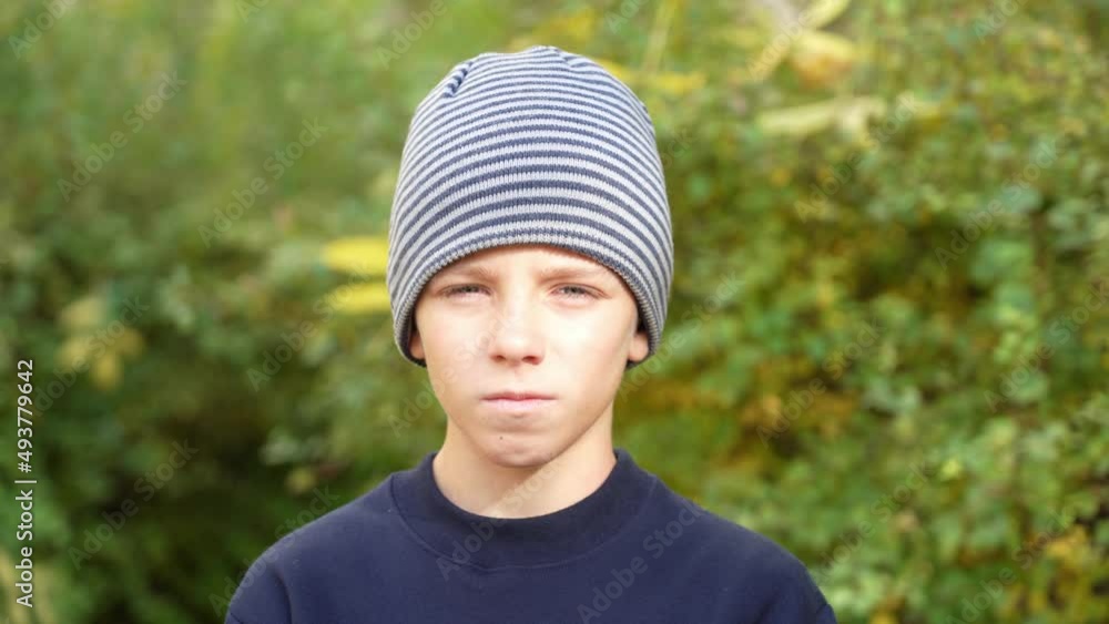 Serious boy in striped hat standing in park looks at camera and nods his head in disagreement. Stylish urban young man leading an external lifestyle. Boy looks around suspiciously at autumn Serious boy in striped hat standing in park looks at camera and nods his head in disagreement. Stylish urban young man leading an external lifestyle. Boy looks around suspiciously at autumn