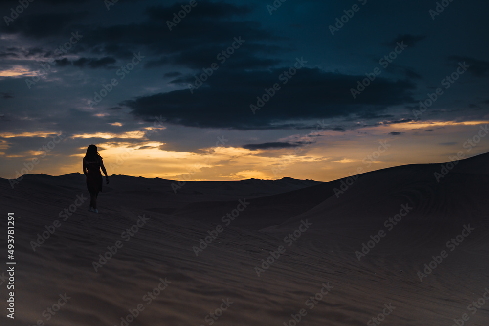 Silhouette of a woman standing alone in the dunes of a desert in sunset time.