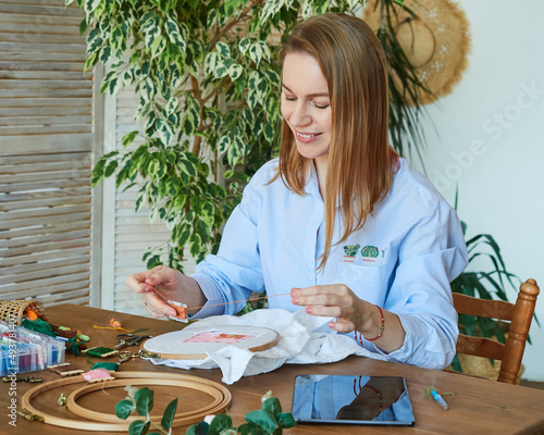 Happy blonde woman cross-stitch, sitting at a wooden table, uses a computer tablet
