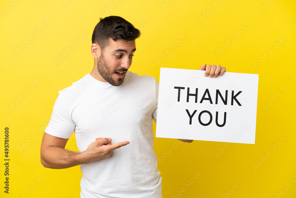 Young handsome caucasian man isolated on yellow background holding a placard with text THANK YOU and pointing it Young handsome caucasian man isolated on yellow background holding a placard with text THANK YOU and pointing it