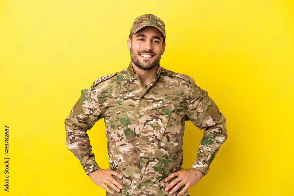 Military man isolated on yellow background posing with arms at hip and smiling Military man isolated on yellow background posing with arms at hip and smiling