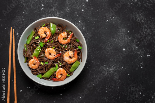 Stir fry buckwheat soba noodles with shrimps and vegetables in a bowl. Black stone background. Asian traditional food. Top view.