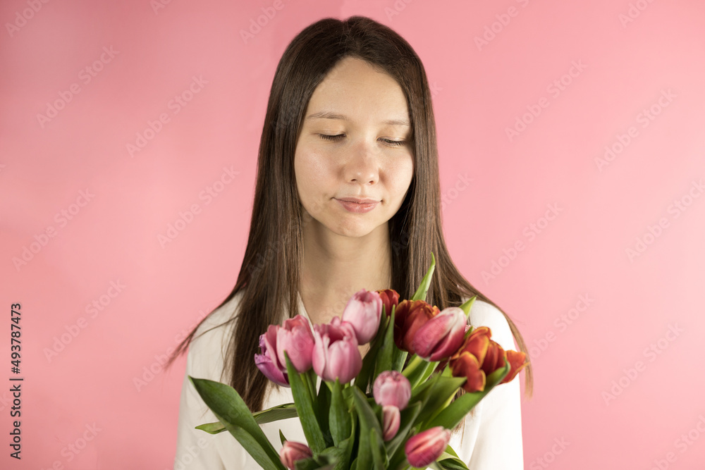 Portrait of a beautiful happy young woman enjoying a bouquet of flowers from tulips, close-up.Delicate pink background.International Women's Day. Spring time. Romance. Softness.Love