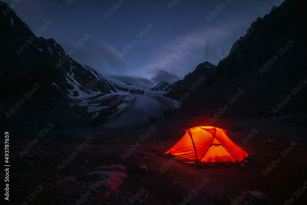 Awesome mountain landscape with vivid orange tent near large glacier tongue under clouds in night starry sky. Tent glow by orange light with view to glacier and mountains silhouettes in starry night.