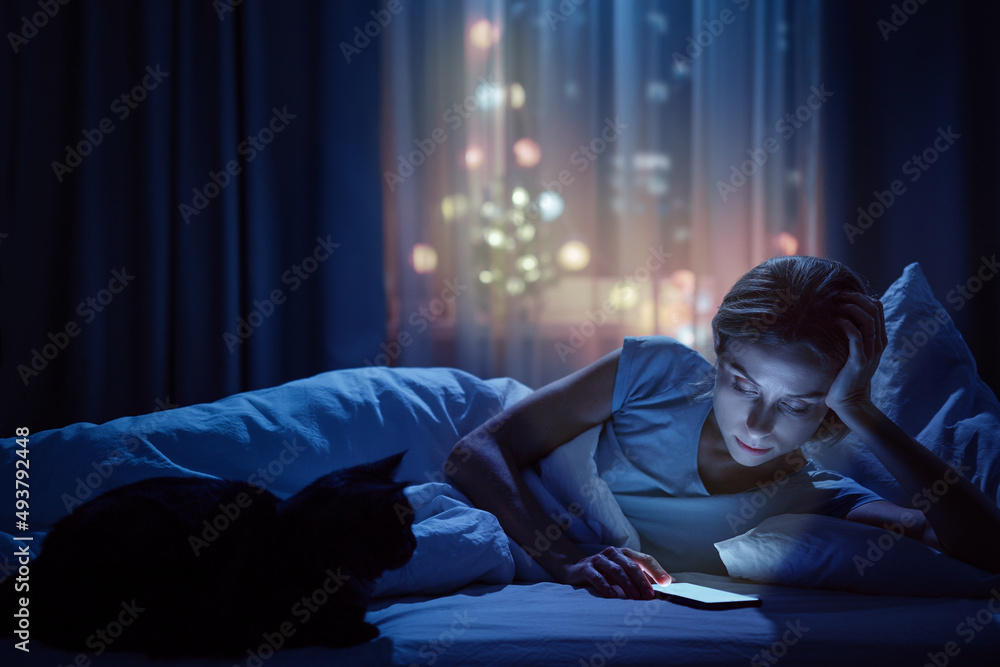 Young woman with her cat using smartphone in bed in her cozy bedroom at night. Young woman with her cat using smartphone in bed in her cozy bedroom at night.