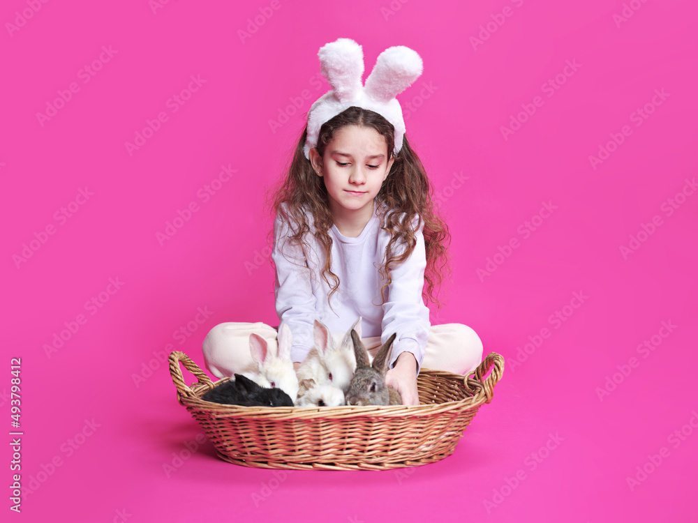 Pretty smiling girl with white bunny ears, showing straw basket with little rabbits, isolated on a pink background.