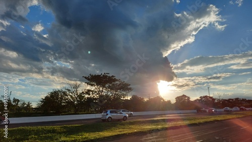 Blue sky, sunset in the road, landscape before dusk, sun between cars and trees, clouds in fantastic shapes and sunbeams between clouds