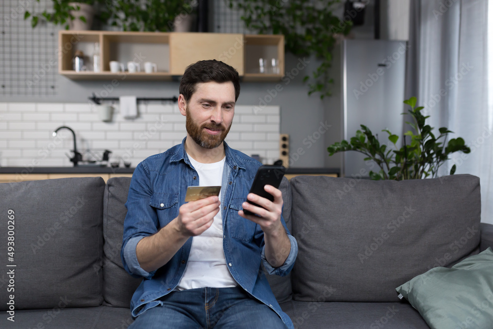 Happy man at home sitting on the couch, shopping in the online store, using the application on the phone, and a bank credit card Happy man at home sitting on the couch, shopping in the online store, using the application on the phone, and a bank credit card