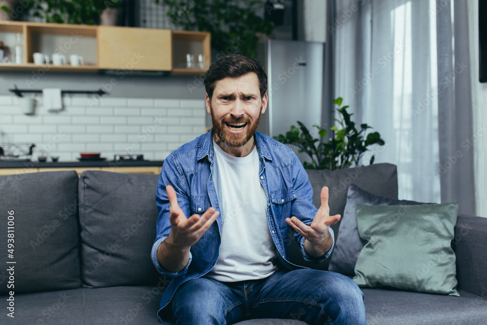 An angry man talking on a video call, looking at the webcam, angrily gesturing with his hands sitting on the couch at home An angry man talking on a video call, looking at the webcam, angrily gesturing with his hands sitting on the couch at home
