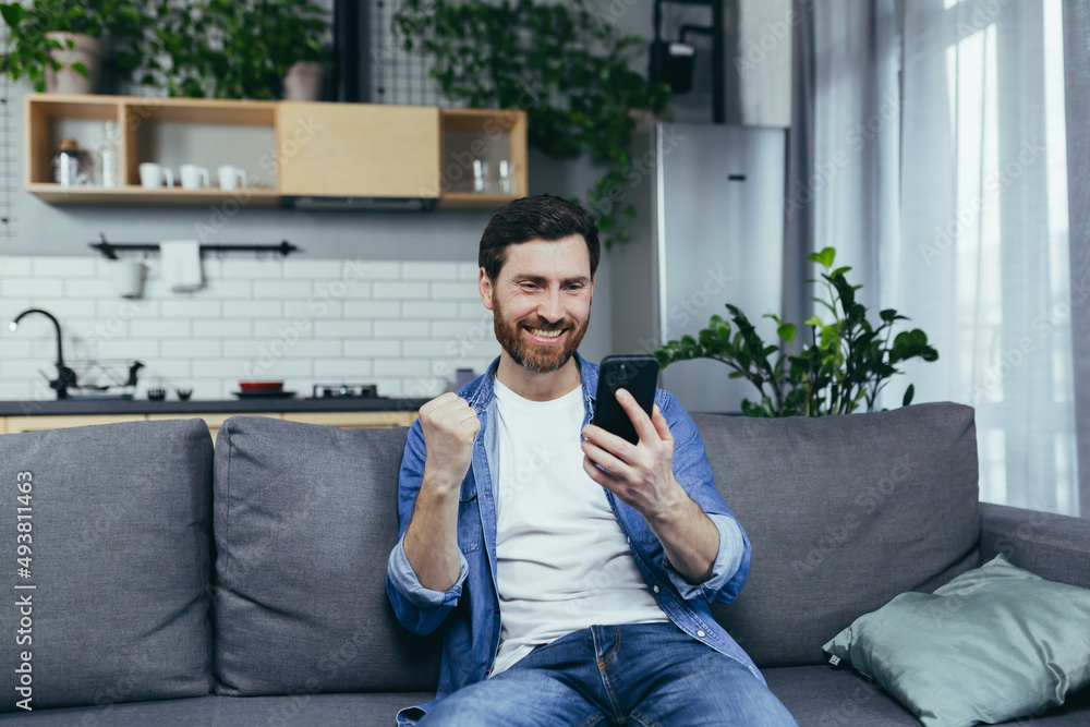 A man at home won and faces the news online, looks at the phone screen and celebrates the triumph of victory, freelancer with phone A man at home won and faces the news online, looks at the phone screen and celebrates the triumph of victory, freelancer with phone