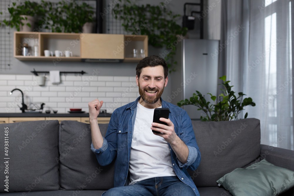 A man at home won and faces the news online, looks at the phone screen and celebrates the triumph of victory, freelancer with phone A man at home won and faces the news online, looks at the phone screen and celebrates the triumph of victory, freelancer with phone