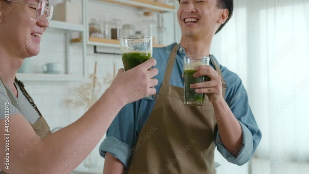 Young smiling gay couple drinking healthy juice with juice machine in the kitchen at home, LGBTQ and diversity concept. Young smiling gay couple drinking healthy juice with juice machine in the kitchen at home, LGBTQ and diversity concept.