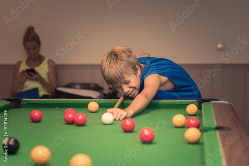 Cute boy in blue t shirt plays billiard or pool in club. Young Kid learns to play snooker. Boy with billiard cue strikes the ball on table. Active Leisure, sport, hobby concept
 photo