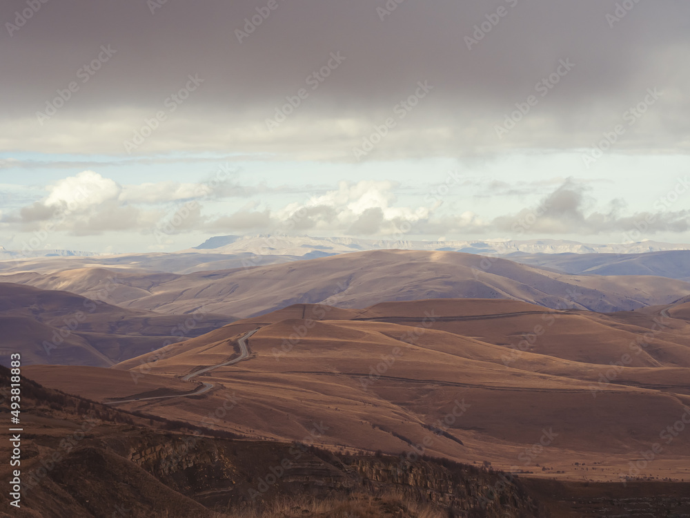 Serpentine road passing through the mountain peaks of the Caucasus with a visible plateau in the distance under the clouds. View from the highlands