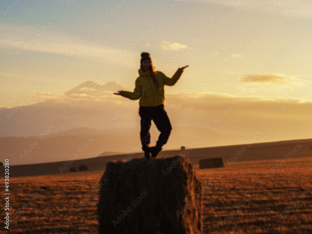 Defocused photo of a woman standing in a pose on a haystack in a wide field against the backdrop of Elbrus