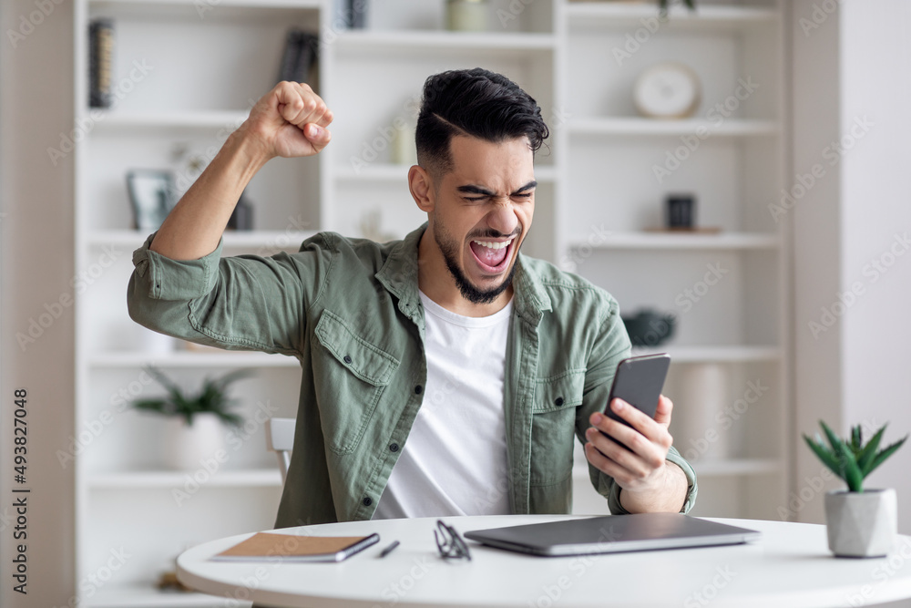 Great News. Overjoyed Young Arab Man Holding Smartphone And Celebrating Success