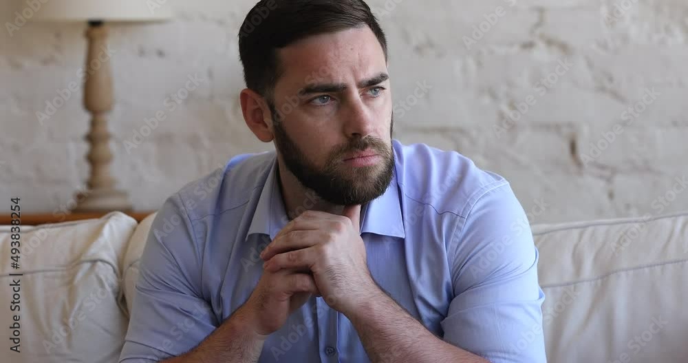 Close up of gloomy pensive millennial man sit on sofa looking depressed, deep in sad thoughts, suffers from mental pain, heartbreak, goes through divorce and break up. Personal life concerns concept