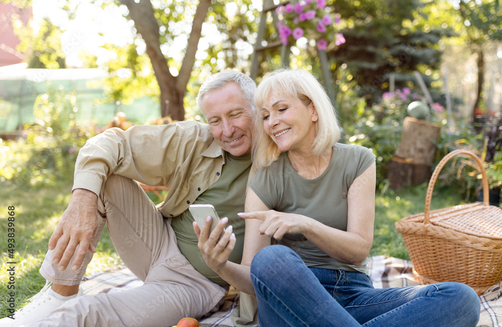 Happy senior spouses sitting on blanket during picnic in garden and using smartphone, resting outdoors on spring day Happy senior spouses sitting on blanket during picnic in garden and using smartphone, resting outdoors on spring day