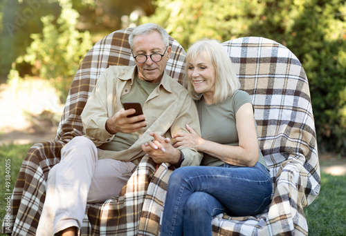 Happy elderly spouses relaxing in wicker chairs and using smartphone, enjoying warm spring evening outdoors © Prostock-studio