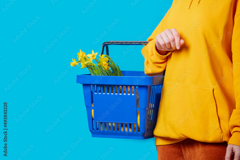 Fototapeta premium Stylish female in yellow hoodie with blue basket and flowers on blue background