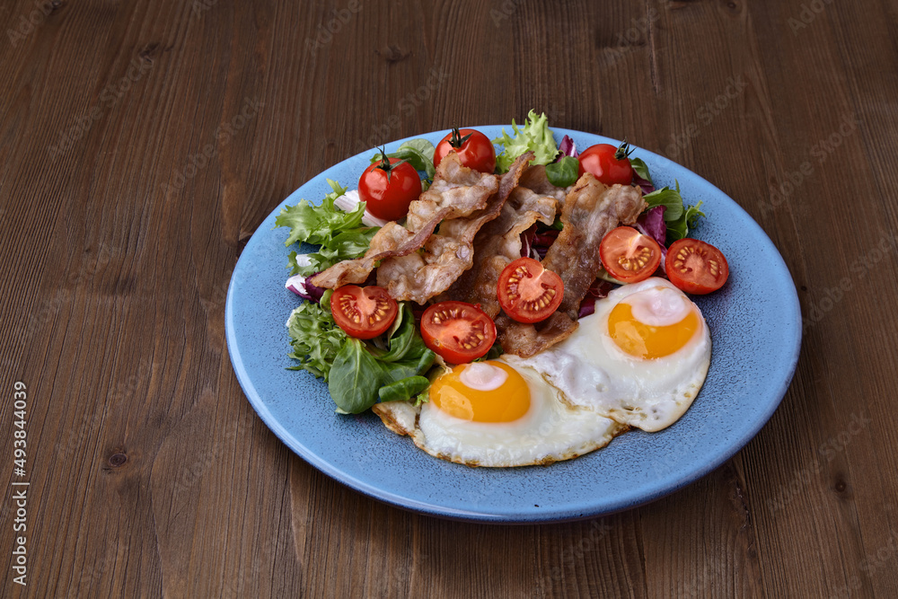 Fried eggs with bacon, lettuce and cherry tomatoes on a blue plate Fried eggs with bacon, lettuce and cherry tomatoes on a blue plate
