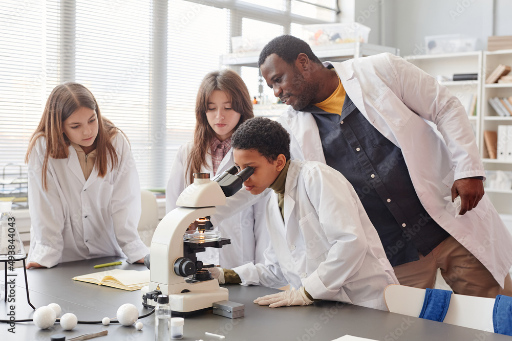 Side view portrait of young black girl looking into microscope with while doing experiments with group of children in school chemistry lab, copy space Side view portrait of young black girl looking into microscope with while doing experiments with group of children in school chemistry lab, copy space