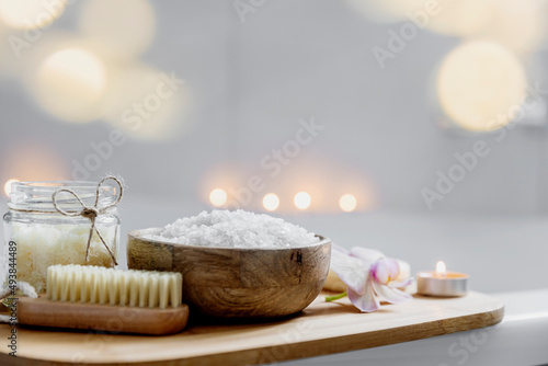 Preparation for hotel spa treatment, home bath procedure. White washbasin in bathroom, accessories on tray. Burning candles, soap, foot brush, towel, glass bottle with sea salt, orchid flower