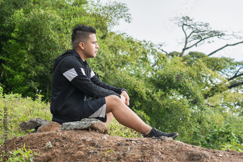 young latin man on top of a colombian mountain, meditating on his life and observing the beautiful nature around him. boy sitting on some rocks after walking along a path. concept of freedom