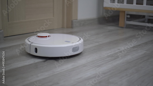 A white robot vacuum cleaner rides on a white parquet floor. The robot with artificial intelligence determines the space and cleans the floor.