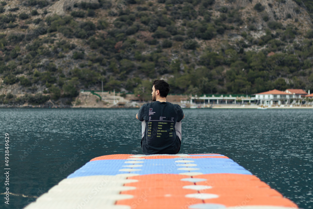 Fototapeta premium Young man sitting alone on dock. He is watching sea view. Her face is not seen in photo. Unrecognizable person. Quiet place.