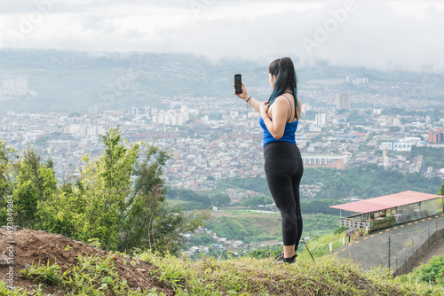 young woman. latin influencer recording a video for her social networks on top of a mountain, in the background the city of Pereira-Colombia. girl with fitnes clothes hiking on top of a hill. photo