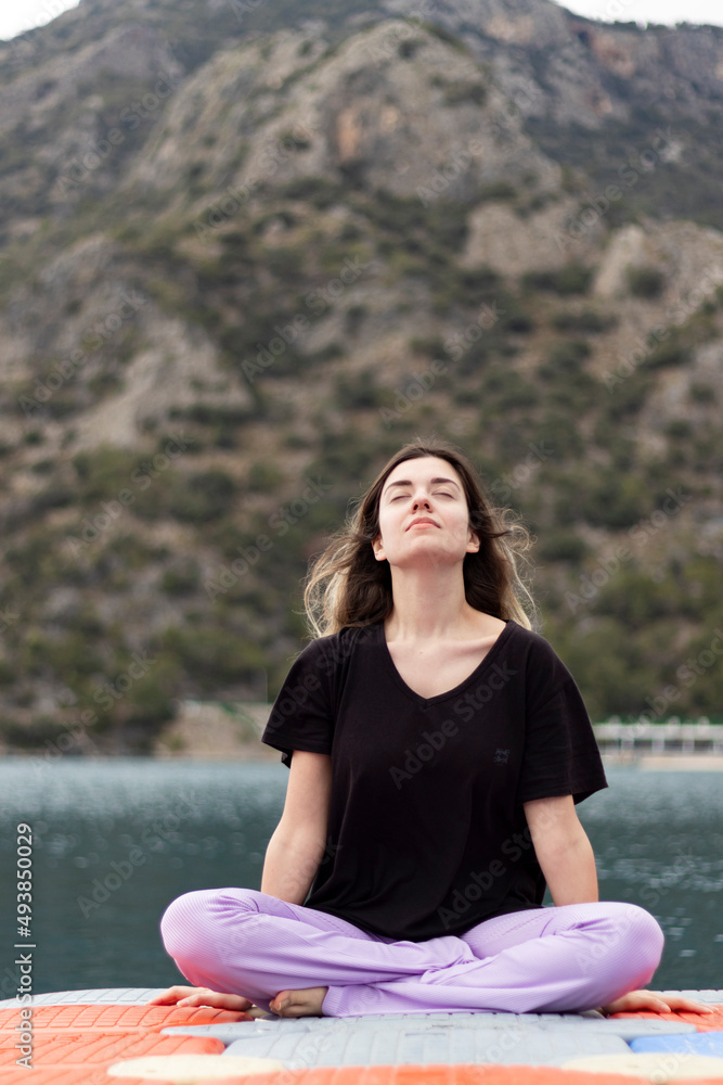 Fototapeta premium Cross-legged young woman. Stretching young girl on colorful dock by sea. Sport, healthy life, meditation and mindfulness. Beautiful landscape. Fresh air concept.