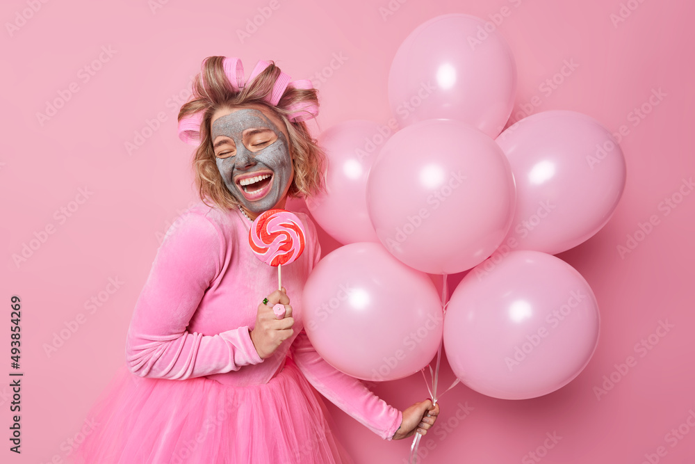 Glad young woman applies clay beauty mask for skin treatment holds caramel candy on stick and bunch of inflated helium balloons wears festive dress makes hairstyle isolated over pink background Glad young woman applies clay beauty mask for skin treatment holds caramel candy on stick and bunch of inflated helium balloons wears festive dress makes hairstyle isolated over pink background