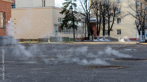 Manhole cover in a paved street on a cold day, as steam escapes from the sewer below up into the city street.