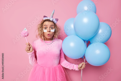 Photo of surprised shocked young woman wears festive dress unicorn headband cannot believe in astonishing news applies beauty patches holds bunch of inflated balloons isolated on pink background © wayhome.studio 