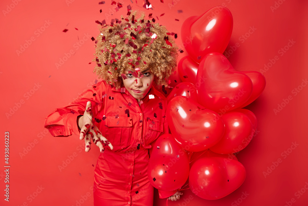 Fototapeta premium Annoyed woman doesnt like surprise received on birthday wears dress holds bunch of heart balloons angry with falling confetti on her has irritated face expression isolated over red background