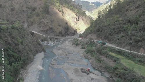 a rocky river bed excavator pulling river rock for cement production bridge spanning over water in valley connecting communities in Kabayan Benguet Philippines wide aerial approach photo