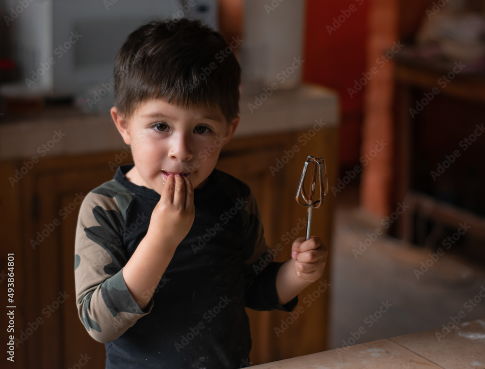 Preschool latin kid looking at camera while tasting raw chocolate cake dough from his hand in the kitchen. Boy holding a whisker. Family lifestyle and joy. Preschool latin kid looking at camera while tasting raw chocolate cake dough from his hand in the kitchen. Boy holding a whisker. Family lifestyle and joy.