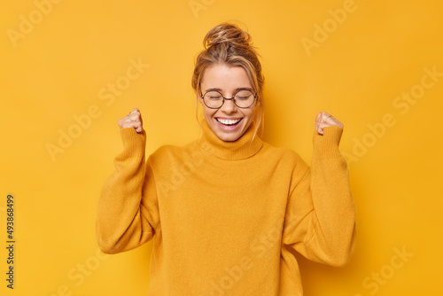 Obraz na plátně Happy positive woman with hair gathered in bun clenches fists rejoices success celebrates victory or great result wears casual jumper round spectacles poses against yellow wall