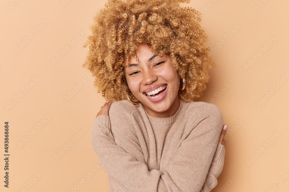 Portrait of happy young woman with curly bushy hair embraces herself feels soft dressed in casual jumper smiles broadly has white teeth isolated over beige background. Self acceptance concept Portrait of happy young woman with curly bushy hair embraces herself feels soft dressed in casual jumper smiles broadly has white teeth isolated over beige background. Self acceptance concept
