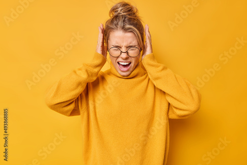 Foto Waist up shot of emotional young blonde woman screams loudly covers ears with hands doesnt want to hear bothering sound wears spectacles and sweater isolated over yellow background