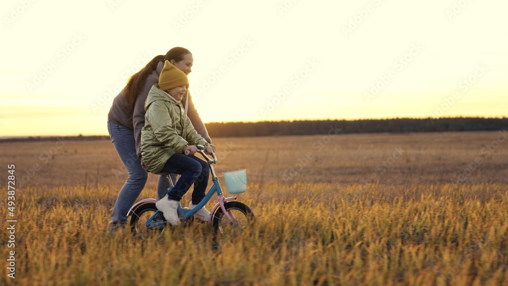 childhood dream. woman mother teaches child to ride bike in the park. Driving lesson for kid from mom at sunset. parent helps child to keep balance and pedal. Kid learns steer bike sun. happy family