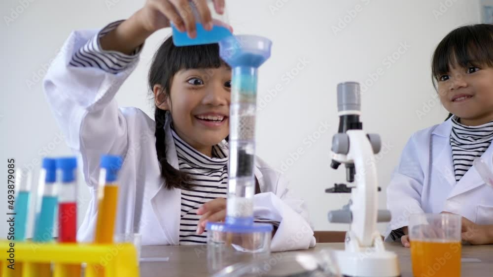 Two Asian siblings wearing coat in lap, little Asian girl pours blue liquid into a water filtration experiment. studying science chemistry with fun Two Asian siblings wearing coat in lap, little Asian girl pours blue liquid into a water filtration experiment. studying science chemistry with fun
