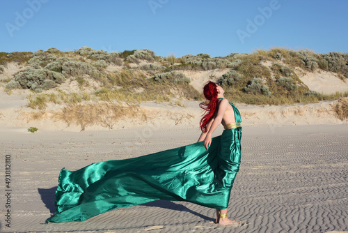 Full length portrait of  red haired woman wearing a  beautiful  long green  silk toga gown. Standing  pose with gestural hands at  ocean beach landscape background.