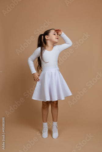 Portrait of young cute smiling girl with long dark hair in white dress, socks and gymnastics shoes standing on toes, looking away with hand over head on brown background. Ballet, ballroom dance.