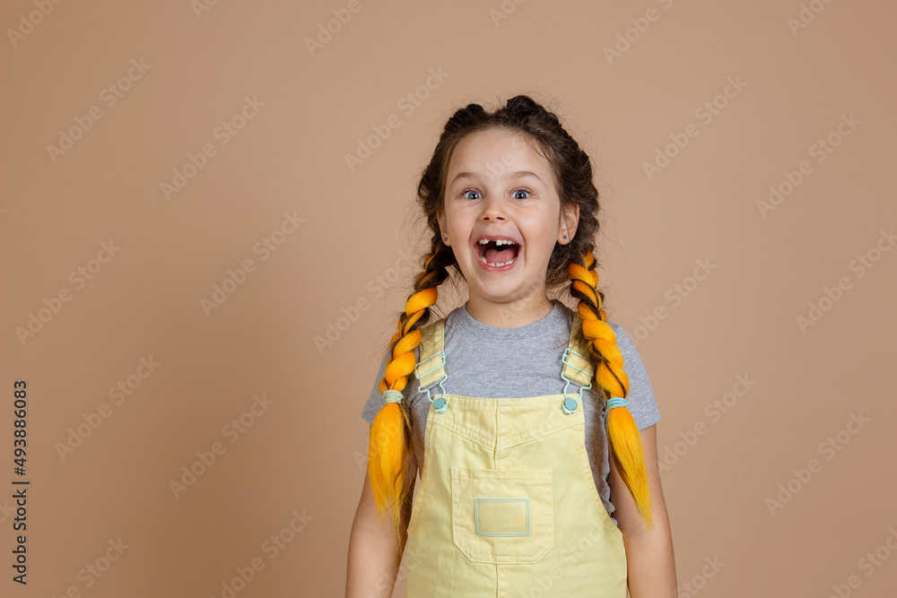 Playful excited small female with yellow kanekalon pigtails, smiling with opened mouth with missing tooth wearing yellow jumpsuit and gray t-shirt on beige background.